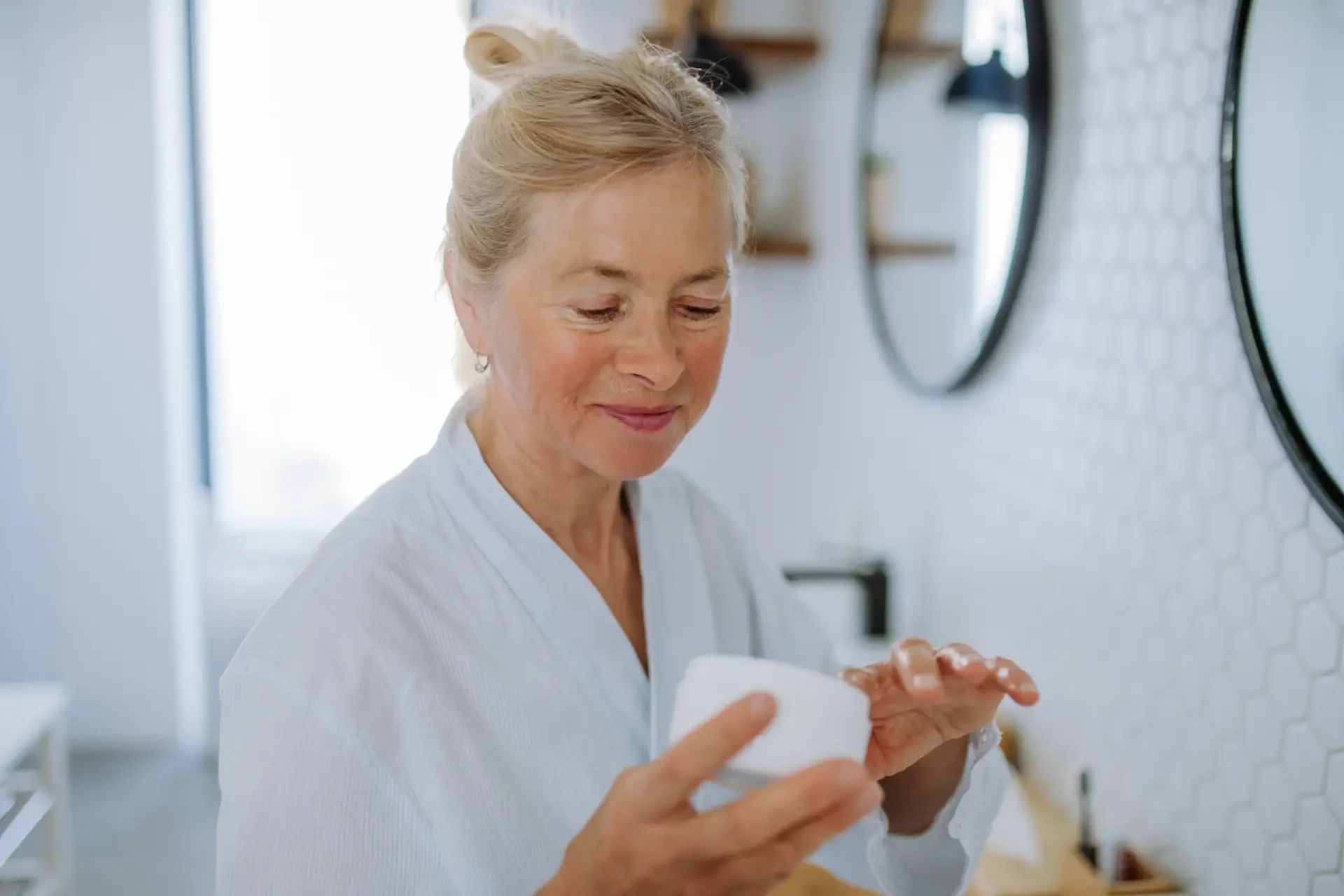 Beautiful senior woman in bathrobe applying natural cream in bathroom, skin care and morning routine